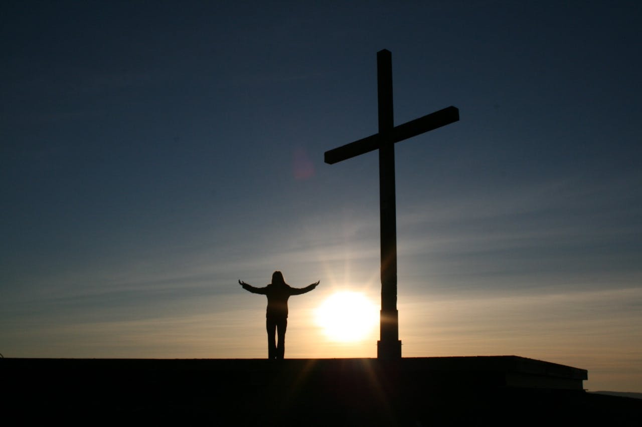 Silhouette of a person with open arms beside a cross at sunset, symbolizing faith and spirituality.